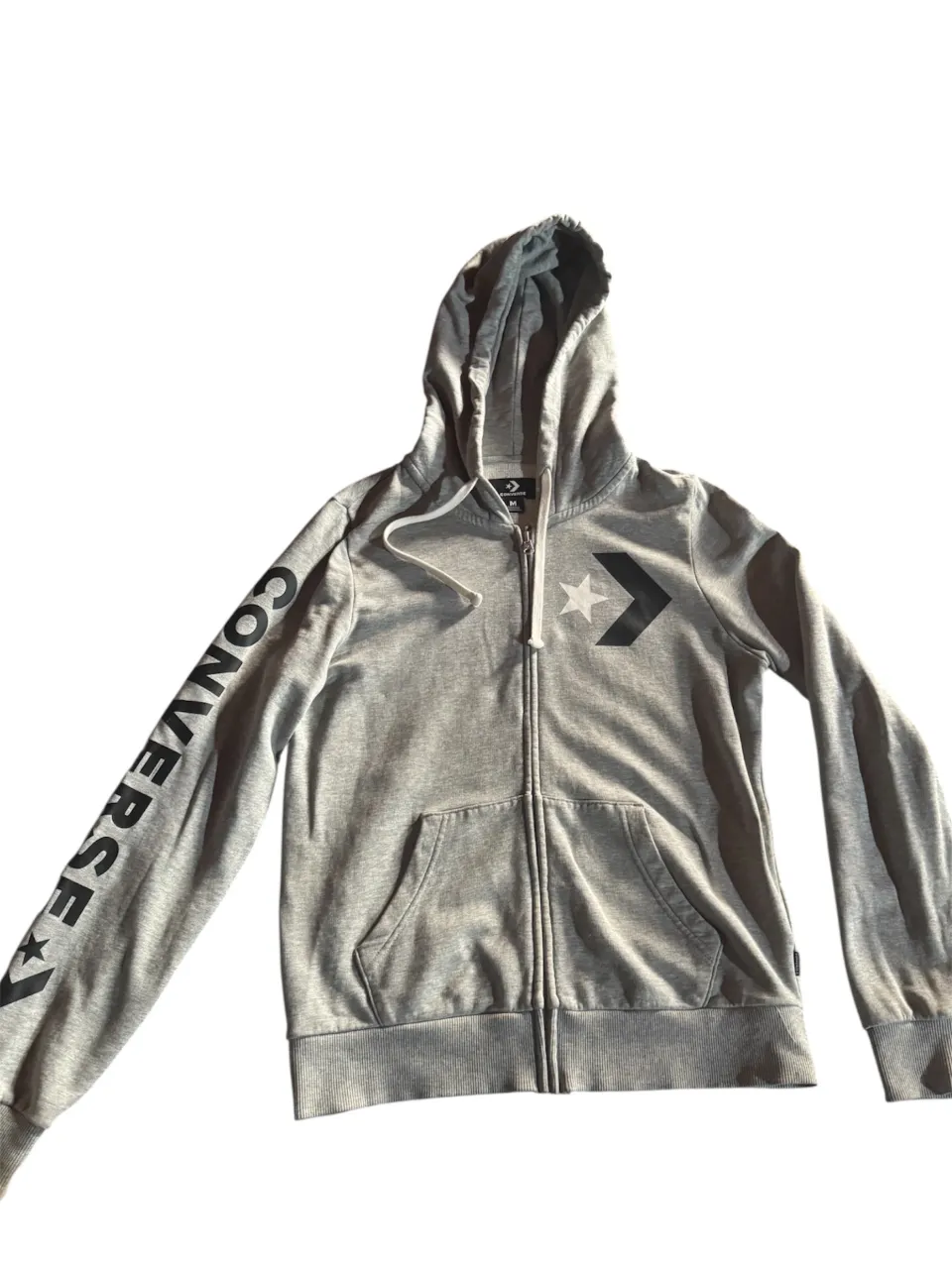 campera gris con capucha y cierre completo, ideal para un look casual y cómodo. Presenta el logo de la marca en el pecho y un estampado de la palabra "CONVERSE" en una de las mangas. Perfecto para el día a día.