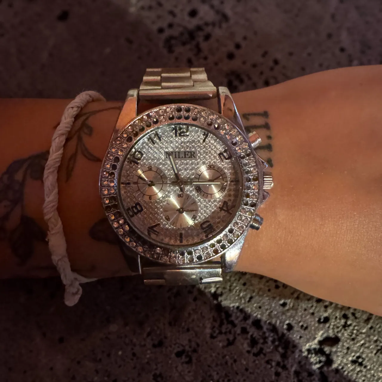 Reloj plateado con detalles de strass en el bisel y en la esfera. Posee tres agujas y tres cronógrafos funcionales. La malla es metálica y ajustable. 