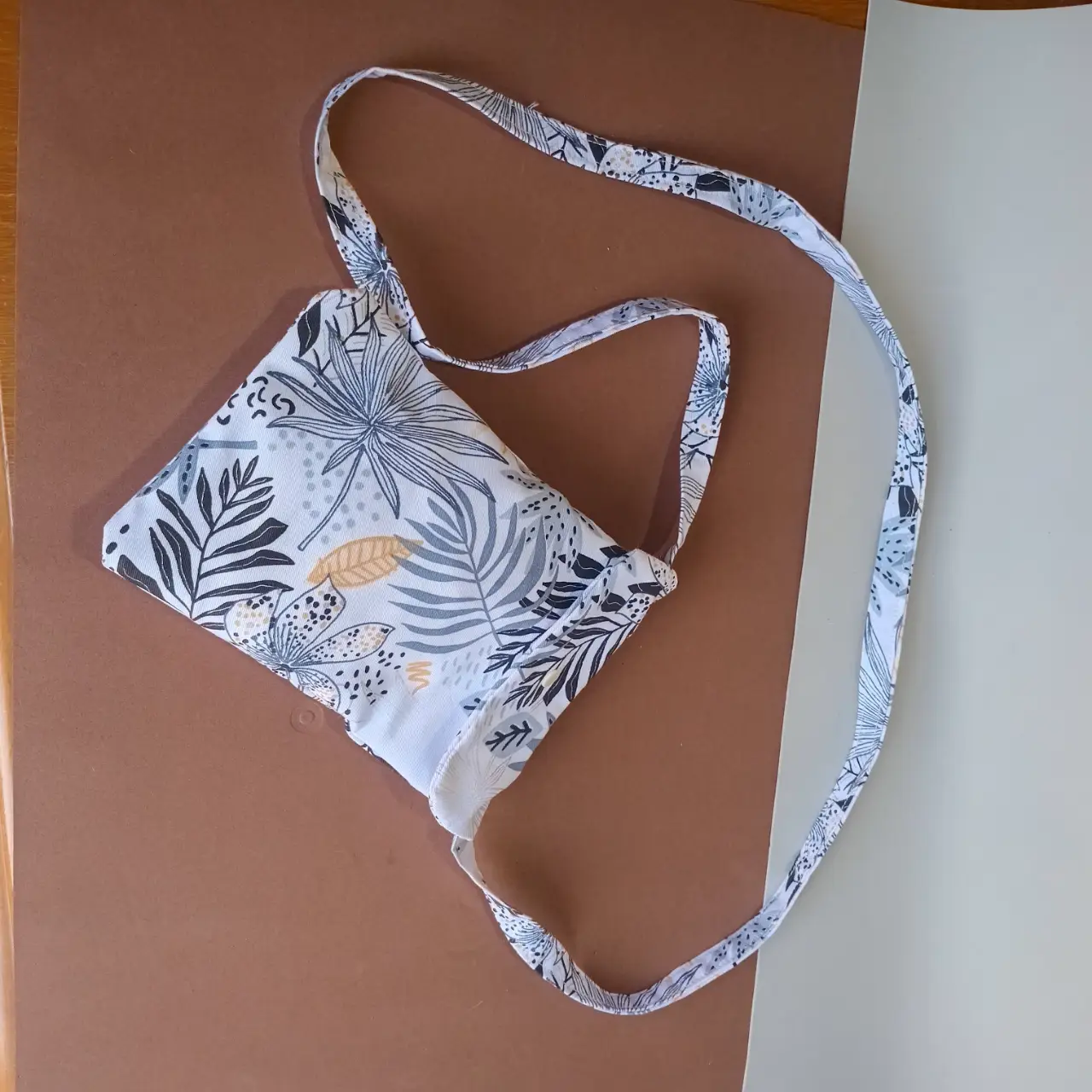 Bolso bandolera con estampado floral en tonos blancos, azules y detalles dorados. Ideal para llevar lo esencial con estilo y un toque artesanal.