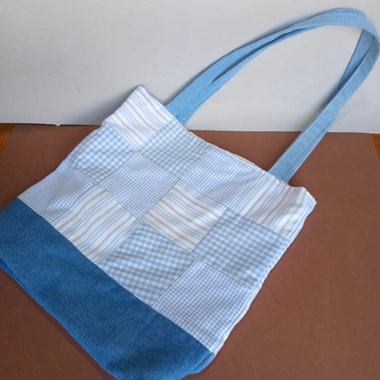 Bolso tote artesanal con diseño patchwork en tonos celeste y blanco. Confeccionado con tela de jean y detalles a cuadros y rayas. Ideal para llevar tus cosas con estilo y originalidad.