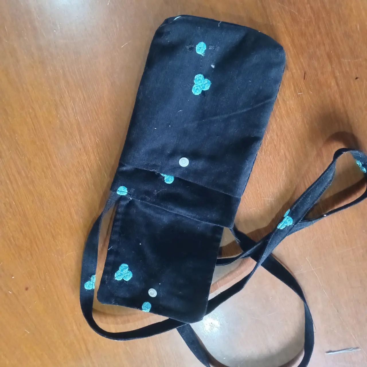 Bandolera negra con detalles bordados en color turquesa y un botón blanco. Ideal para llevar lo esencial con estilo y comodidad.