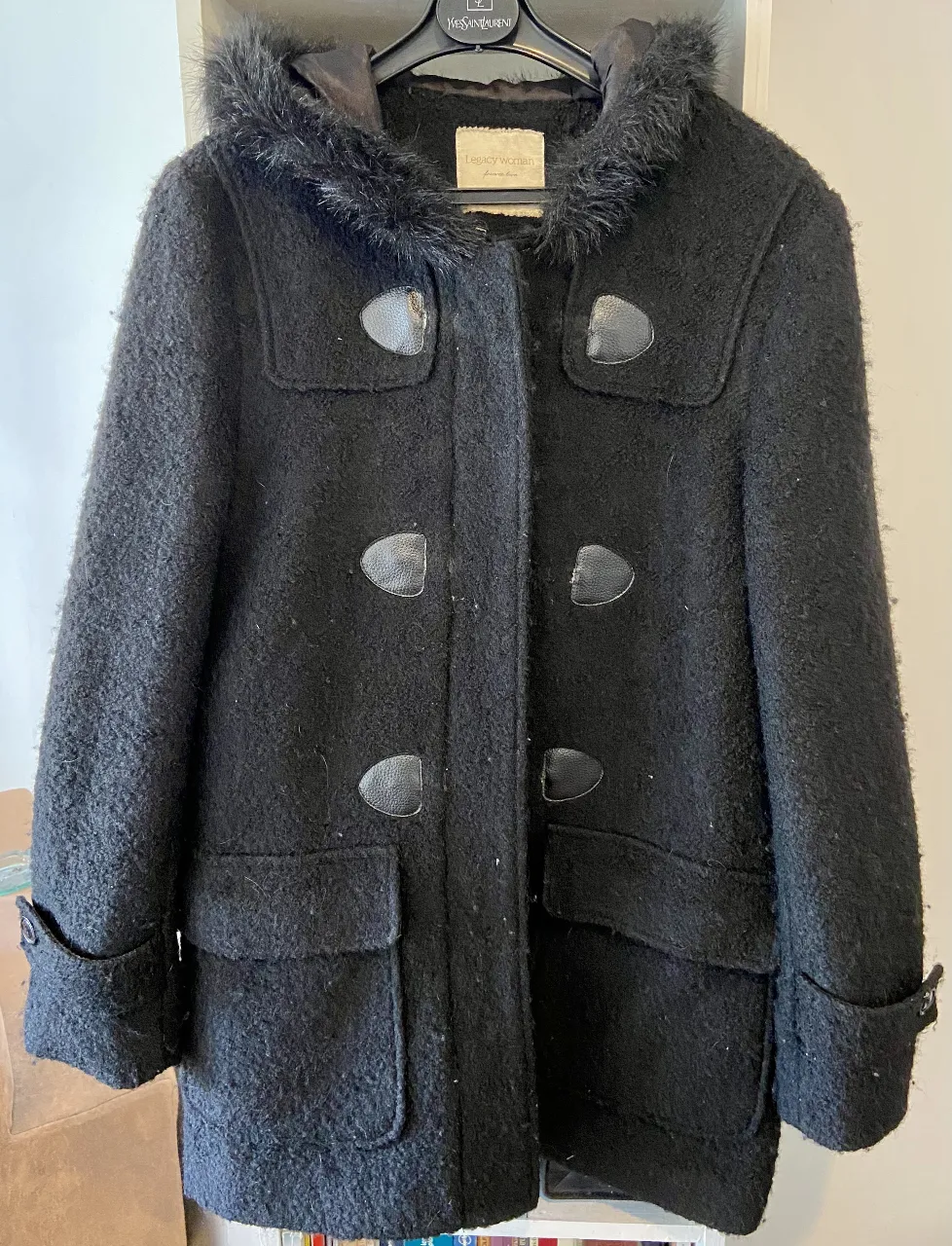 Abrigo tipo "duffle coat" negro, con capucha forrada en piel sintética y detalles de cuero en los cierres. Posee bolsillos frontales y un diseño clásico ideal para el frío.