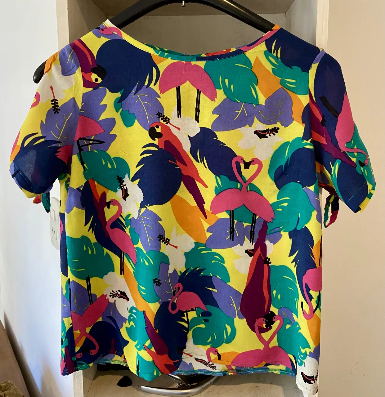 Remera súper colorida y alegre con estampa de flamencos y loros. Ideal para darle un toque divertido a tu look de verano. Tiene mangas cortas con lazos y un detalle de corte en la espalda.