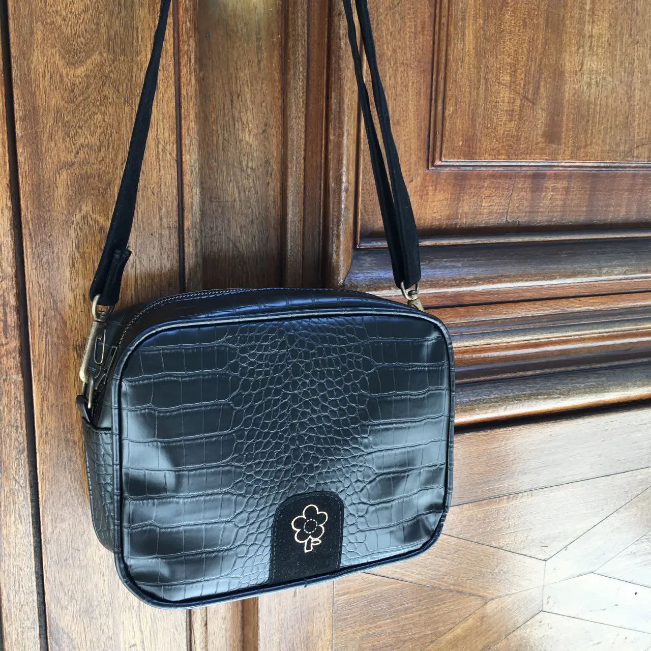 Bandolera negra con textura de cocodrilo y detalle de flor en la solapa. Ideal para un look elegante y moderno.