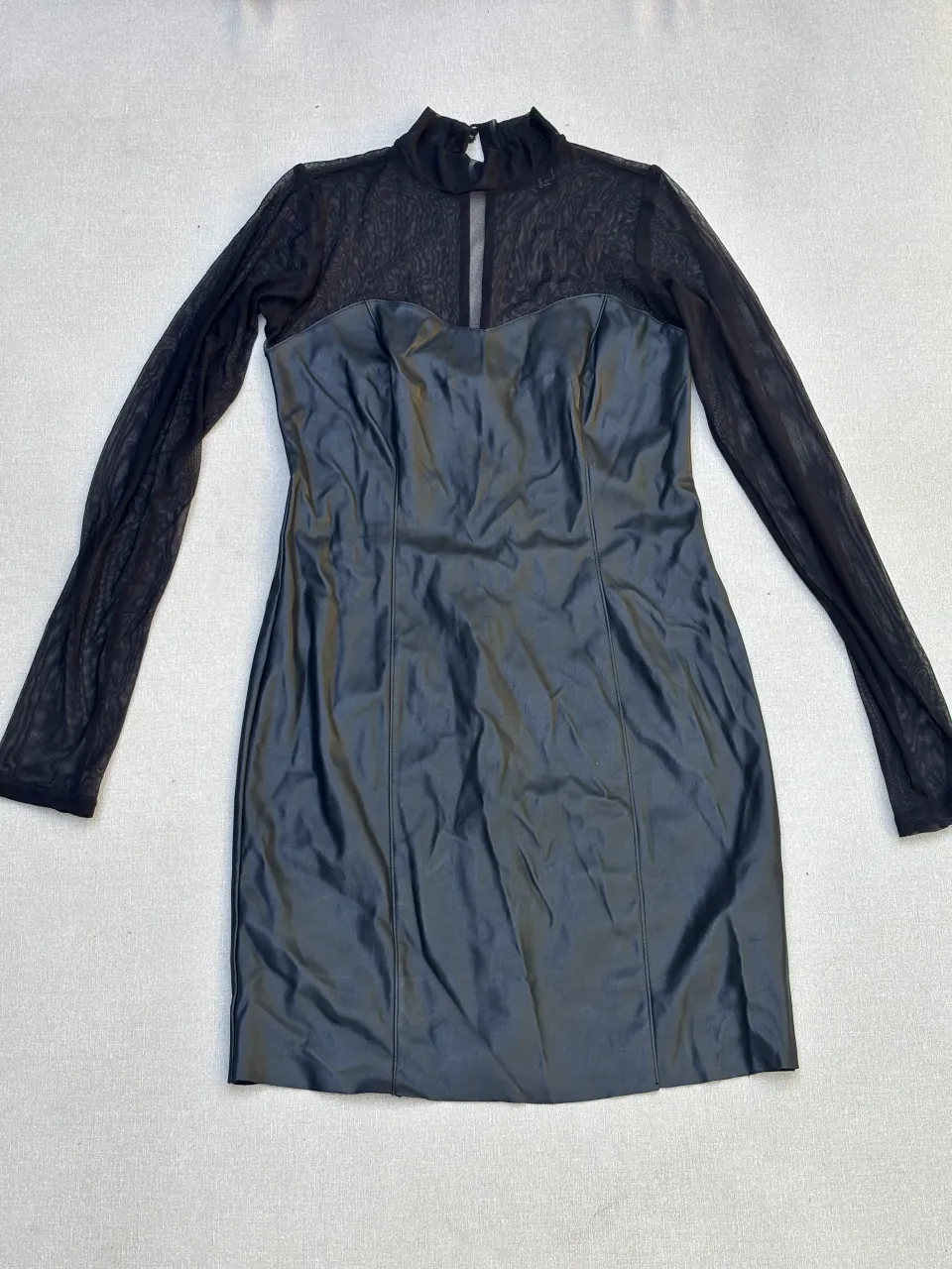 Vestido negro de cuero sintético con mangas largas de tul. Diseño ajustado al cuerpo con cuello alto y detalles de transparencia en el escote. Ideal para una salida nocturna o una ocasión especial.