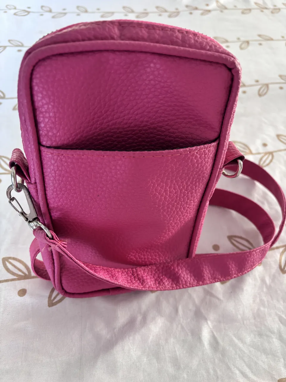 Cartera pequeña de color fucsia con correa larga ajustable. Posee un bolsillo exterior abierto y un compartimento principal con cierre.