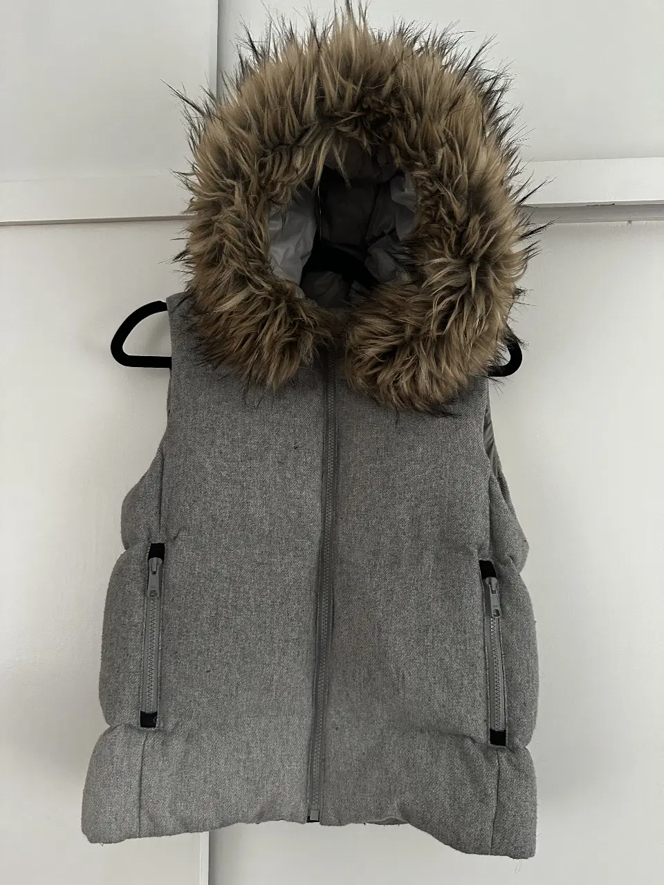 Chaleco de abrigo gris con capucha de piel sintética. Ideal para el invierno, con cierre y bolsillos con cremallera. Perfecto para un look casual y abrigado.