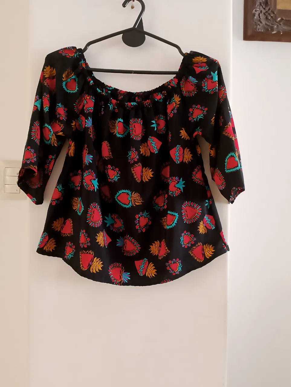 Blusa de mujer con estampado de corazones rojos y detalles en turquesa y dorado sobre fondo negro. Diseño off-shoulder con mangas 3/4, ideal para un look bohemio y casual.