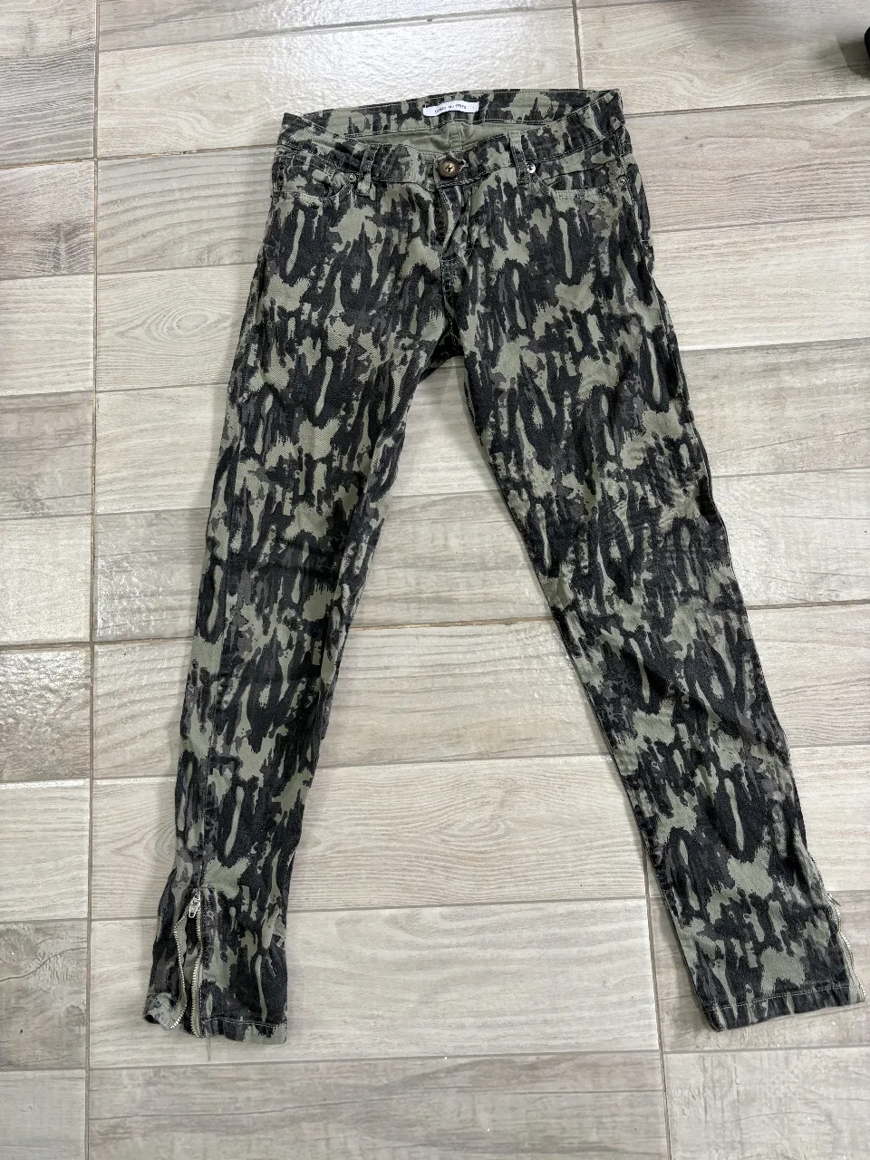 Pantalón de jean recto verde militan con estampado camuflado en tonos grises y negros. Con cierres en los tobillos.