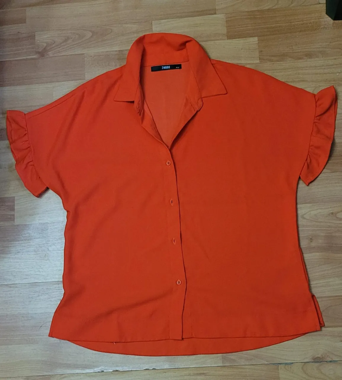 Blusa naranja con mangas cortas y volados en los hombros. Cuello camisero y botones frontales. Ideal para un look casual y moderno.