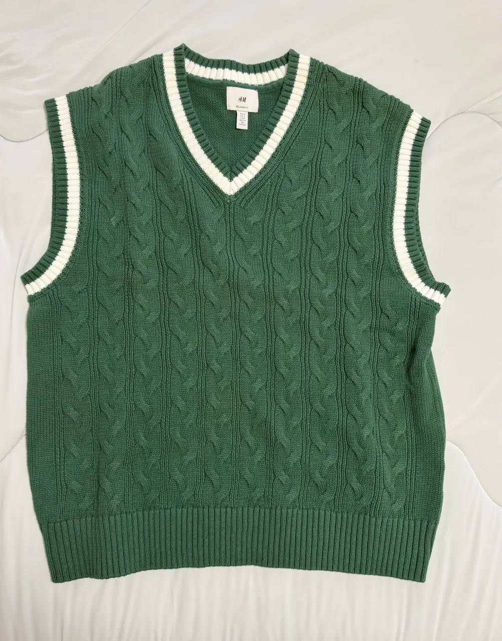 Chaleco de tejido verde con cuello en V y detalles de trencitas. Posee un borde blanco en el cuello y en los hombros.