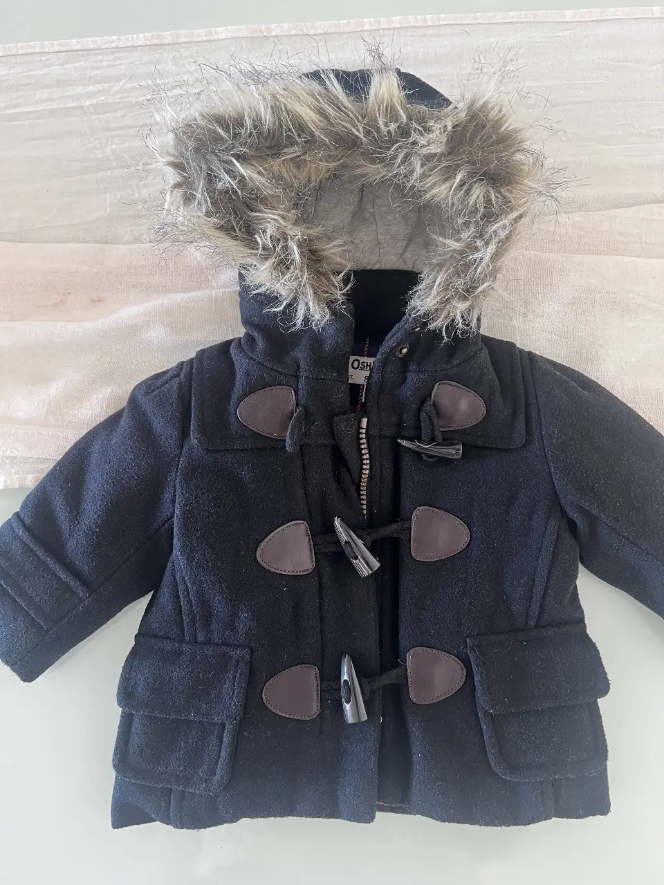 Campera de abrigo para niño, estilo duffle coat, con capucha con borde de piel sintética. Cierre frontal con cremallera y broches de madera. Interior forrado con tela escocesa roja y negra. Ideal para el invierno.