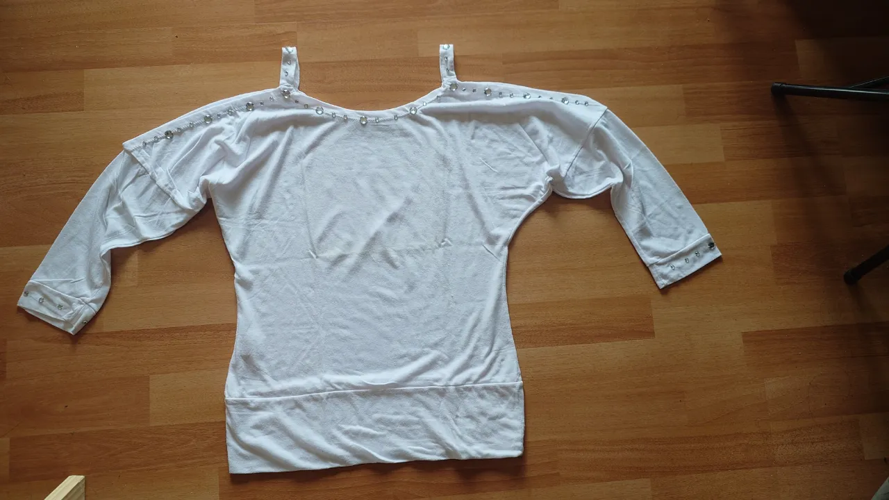 Blusa blanca de manga 3/4 con detalles de strass en el cuello y hombros. Diseño con hombros descubiertos y detalle de volado en la parte superior. Ideal para un look casual y femenino.