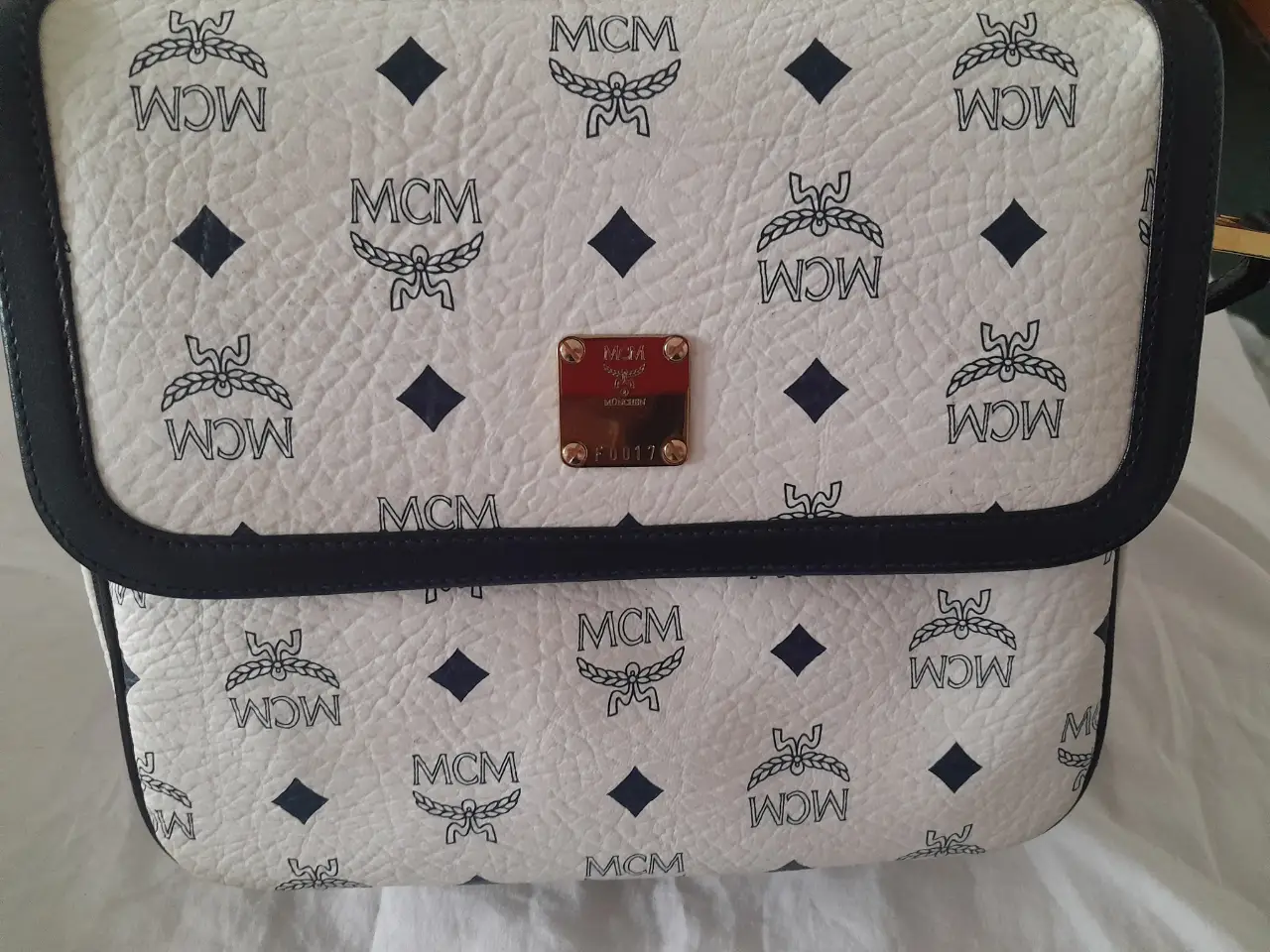 Cartera bandolera MCM en cuero blanco con estampado del logo de la marca y detalles en negro. Interior con cierre y detalles dorados. Ideal para un look casual y elegante.inglesa