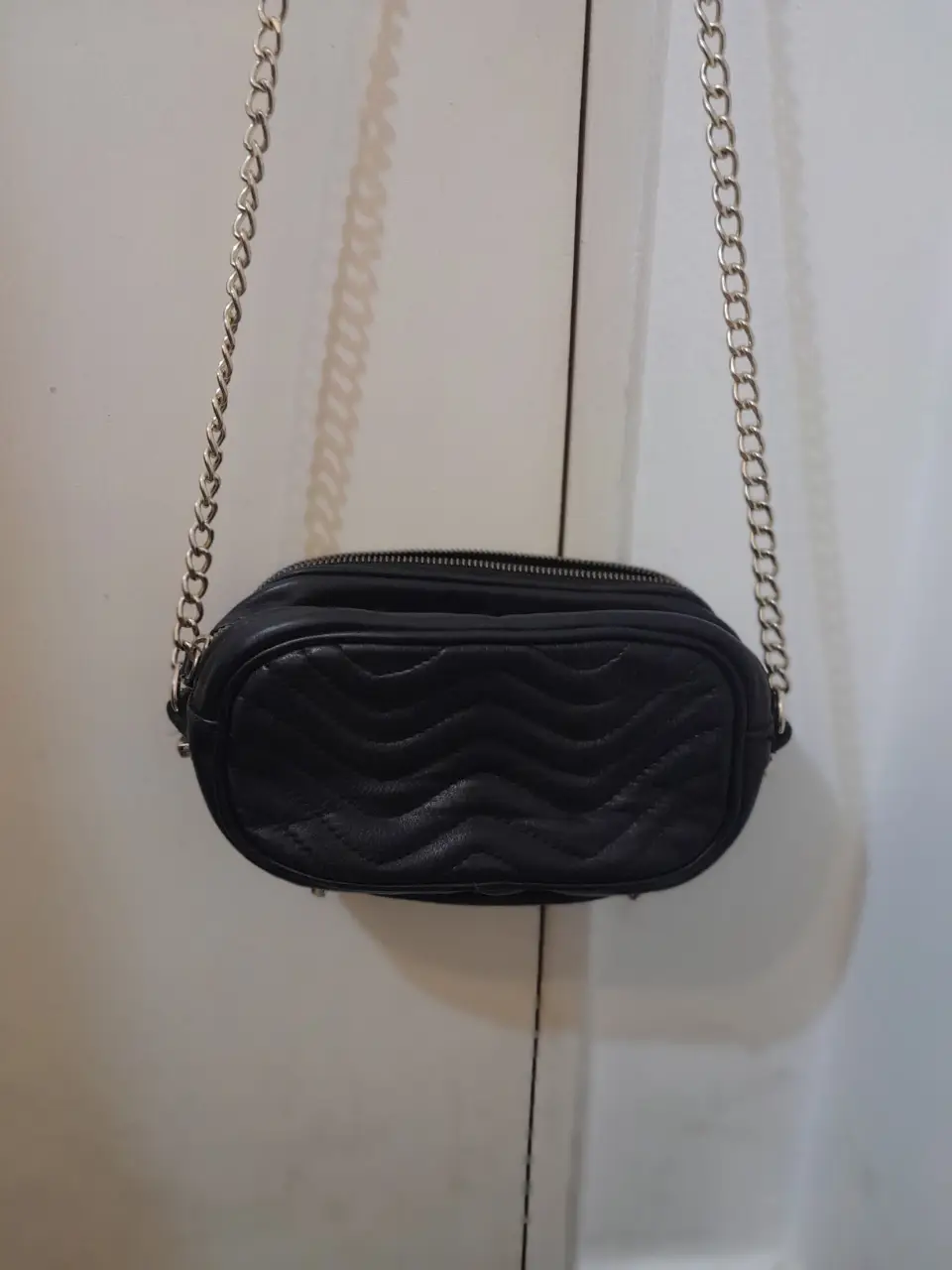Cartera bandolera negra con diseño acolchado en forma de zigzag y cadena dorada. Ideal para un look moderno y versátil.