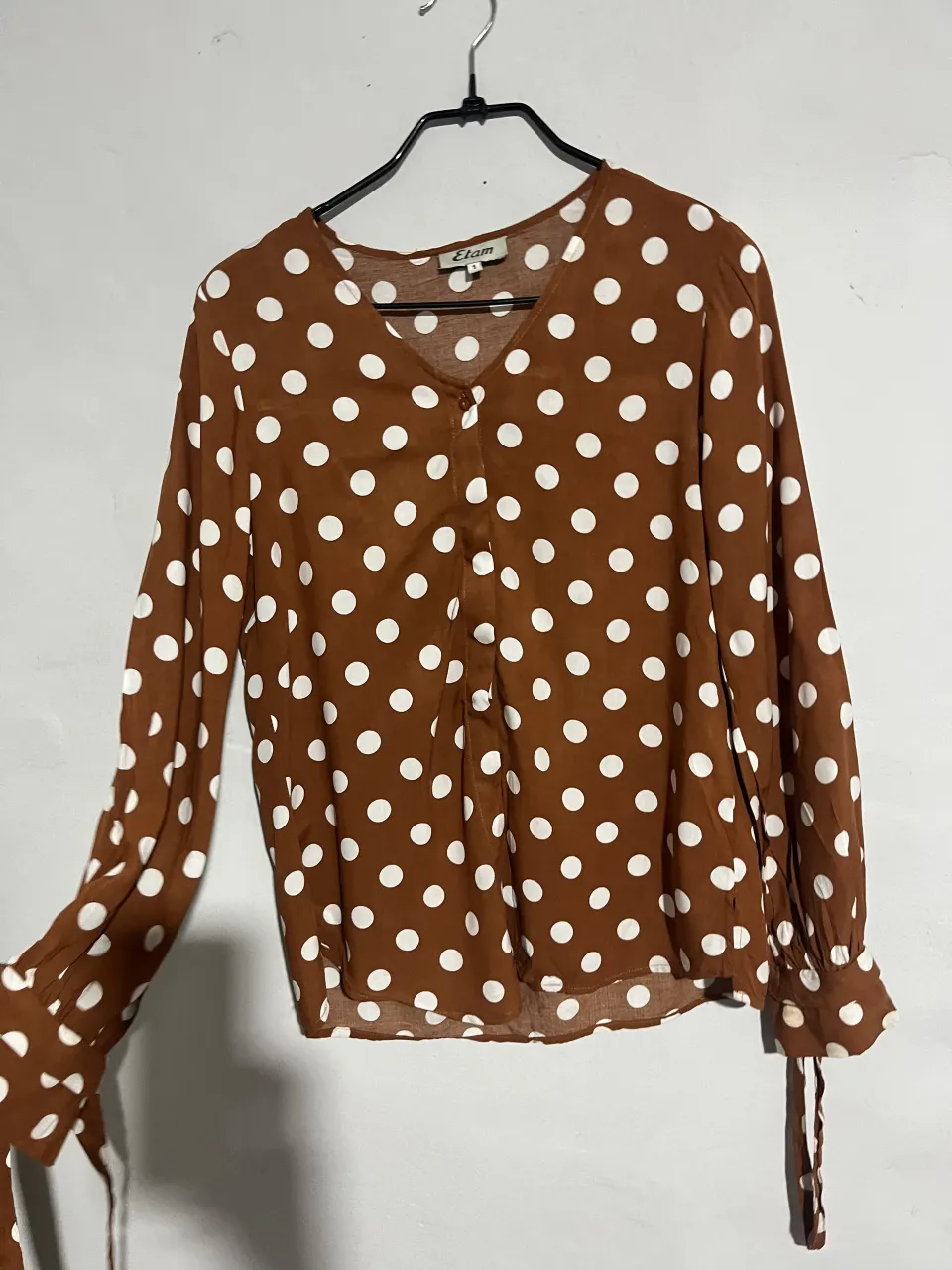 Blusa marrón con lunares blancos, manga larga y escote en V. Detalle de lazo en los puños. Ideal para un look casual o elegante.