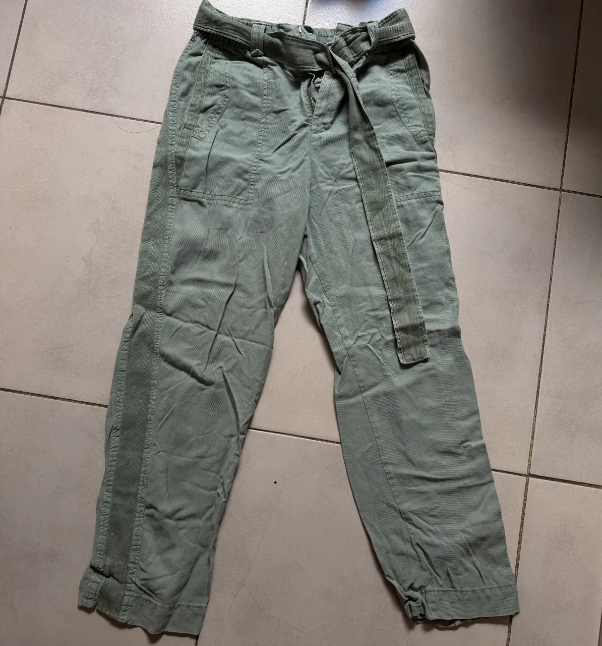 Pantalón de color verde militar con cinturón de tela anudado en la cintura. Posee bolsillos frontales y detalles de costura a lo largo de las piernas.