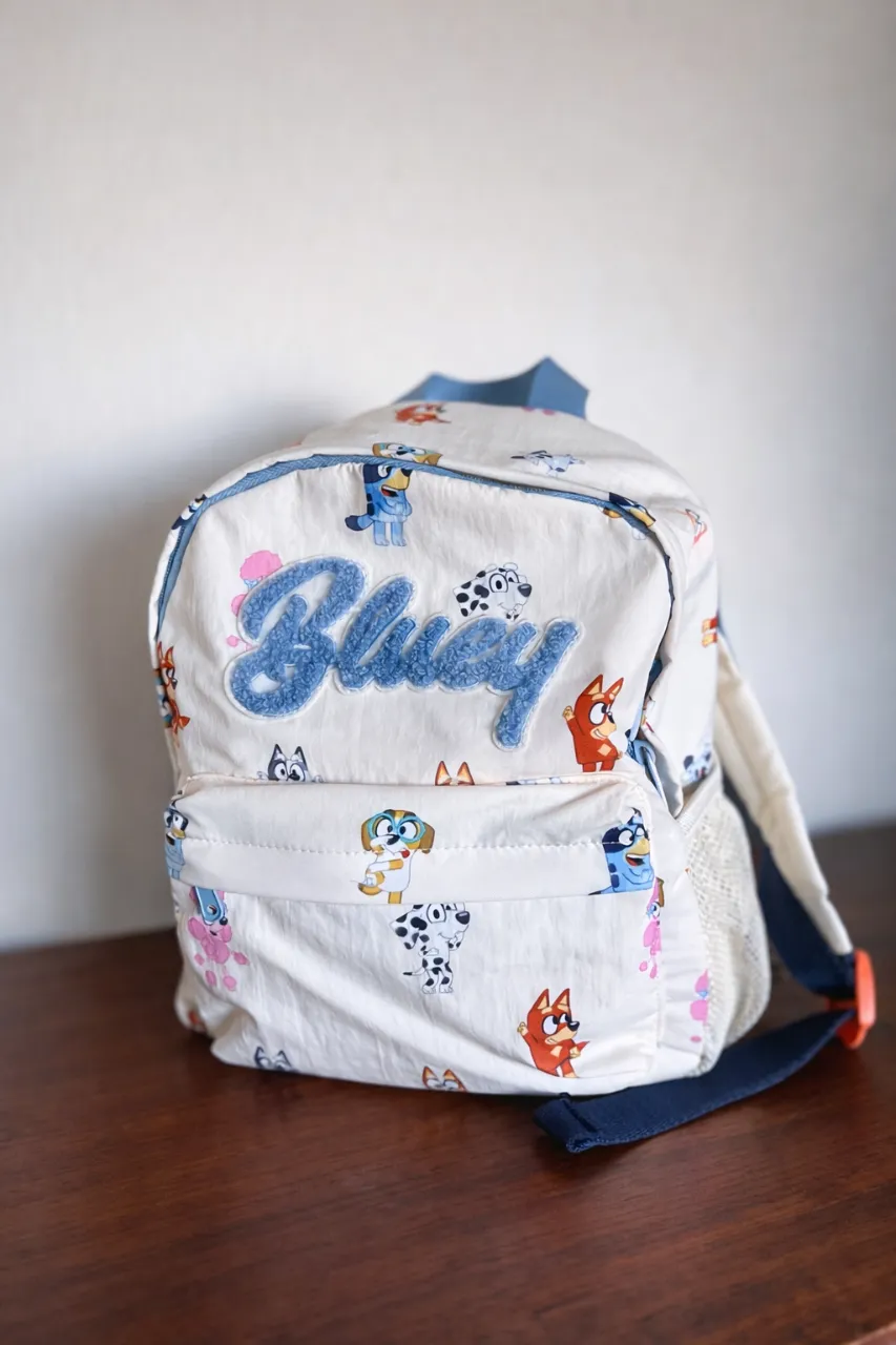 Mochila infantil con estampado de personajes de Bluey. Tiene un bolsillo frontal con cierre y bolsillos laterales de red. Las tiras son regulables y acolchadas.