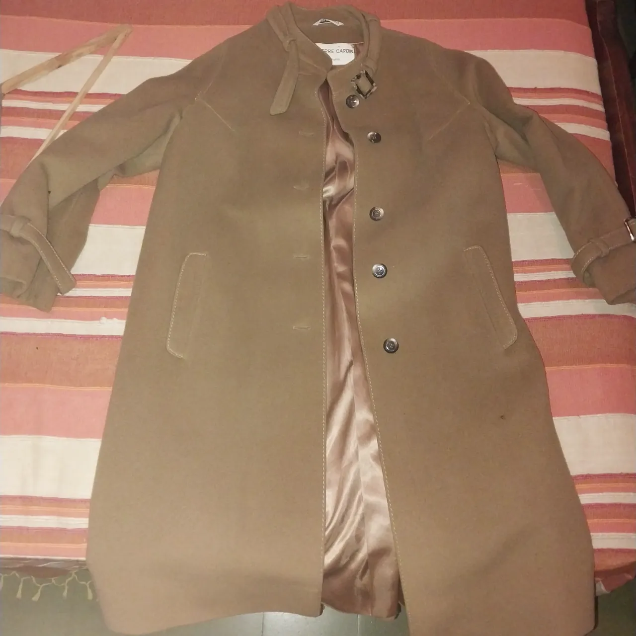 Tapado de paño color marrón. Tiene cuello con solapa y cierre de botones al frente. Los puños de las mangas son anchos y tienen un detalle de hebilla. Posee bolsillos laterales.