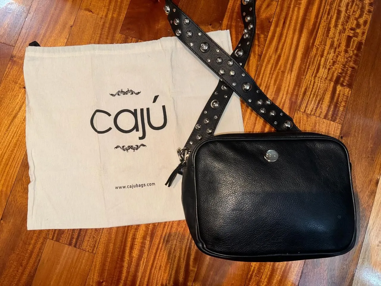 Cartera bandolera de cuero negro con correa ajustable y detalles de tachas. Incluye bolsa protectora con logo. Ideal para un look casual y moderno.