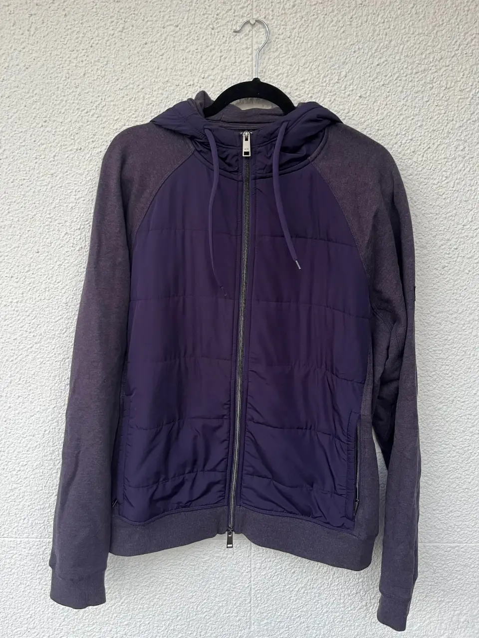 Buzo con capucha para hombre, con cierre frontal y paneles acolchados en color violeta. Mangas y capucha en gris oscuro más tirando a violeta clarito. Ideal para un look casual y abrigado. 
