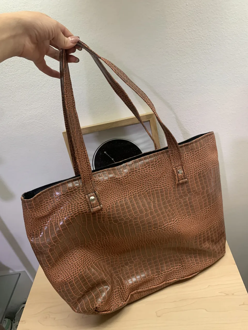 Bolso tote de cuero sintético con textura de cocodrilo en color marrón. Ideal para el día a día, tiene un amplio compartimento principal y un bolsillo interior para organizar tus pertenencias. Sus asas son cómodas para llevar al hombro. Un accesorio versátil que combina con todo.