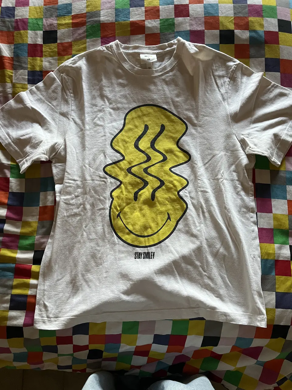 Remera blanca de algodón con estampado de un smiley amarillo distorsionado. Estilo casual y moderno, ideal para un look urbano.