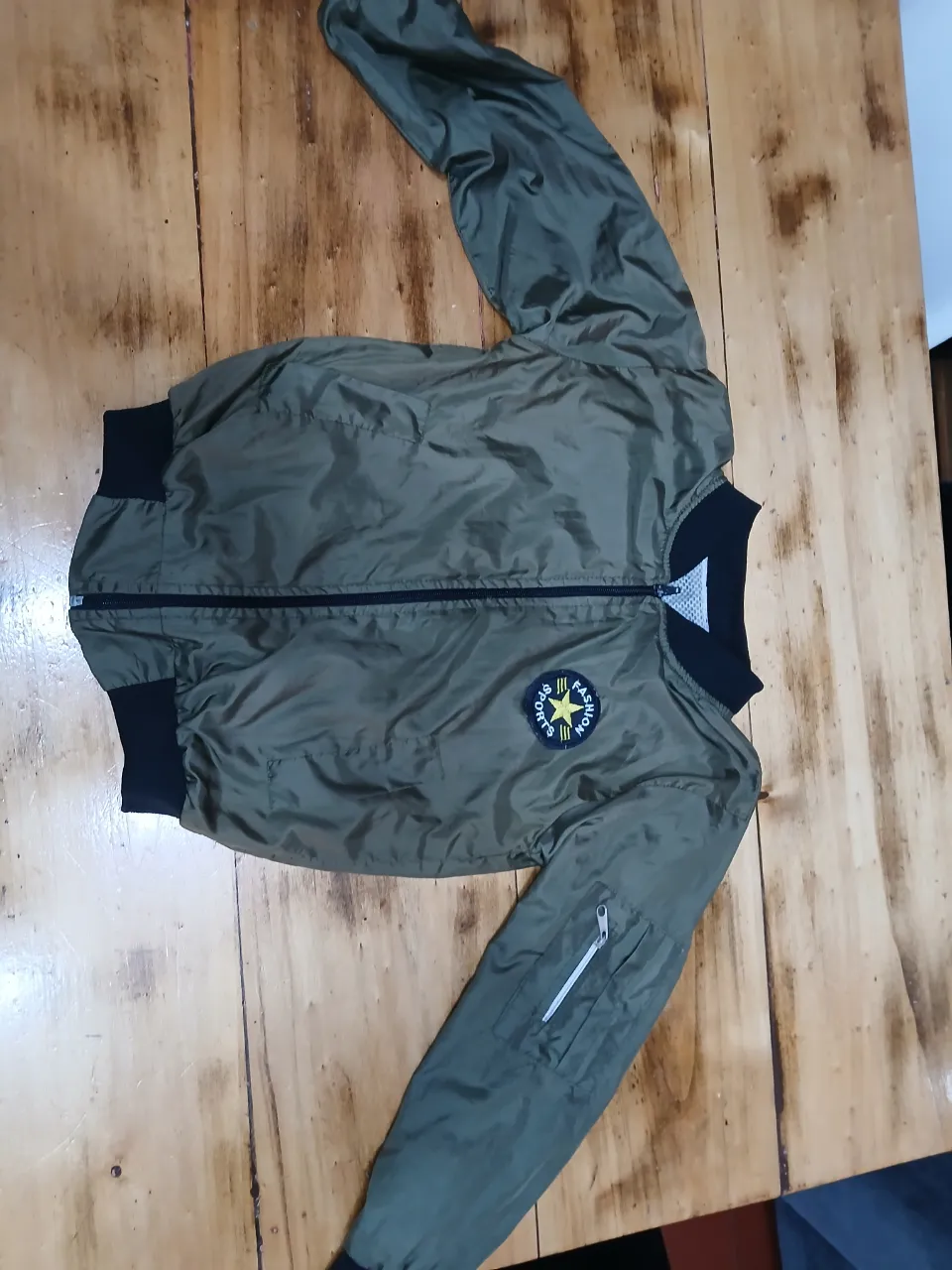 Campera bomber verde militar con detalles en negro. Posee un parche en el pecho y un bolsillo con cierre en la manga. Ideal para un look casual y canchero.