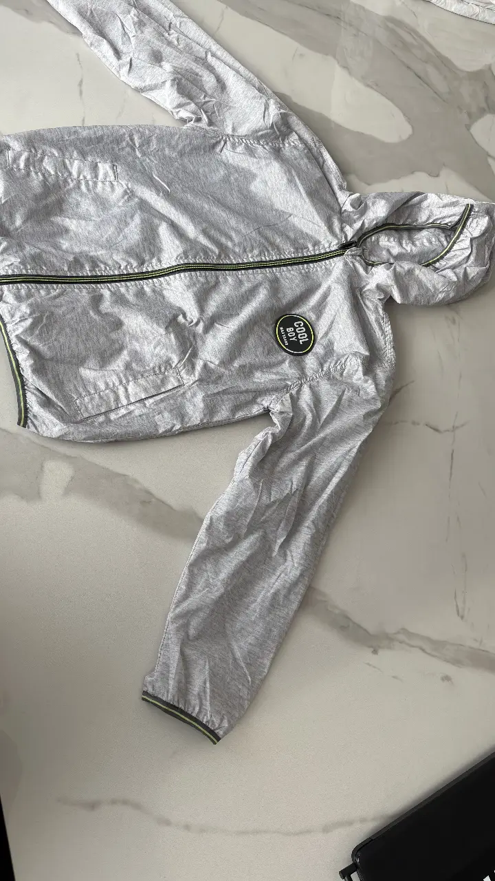 Campera rompe viento gris con capucha para niño. Detalles en verde y cierre. Logo 'Cool Boy' en el pecho. Ideal para actividades al aire libre. 