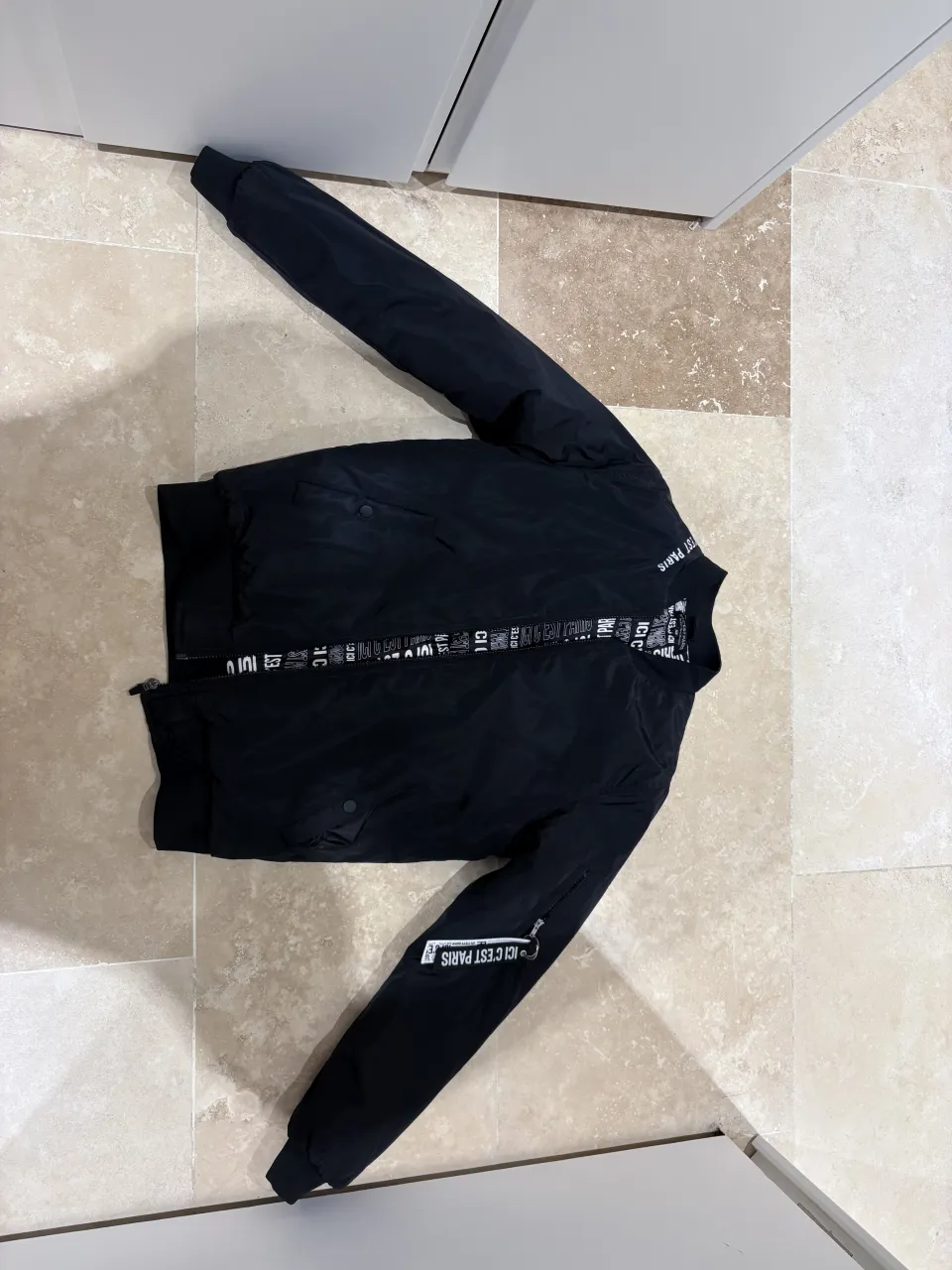 Campera bomber negra con inscripciones en blanco. Tiene cierre al frente y un bolsillo con cierre en la manga. El forro interior está estampado con el mismo diseño.