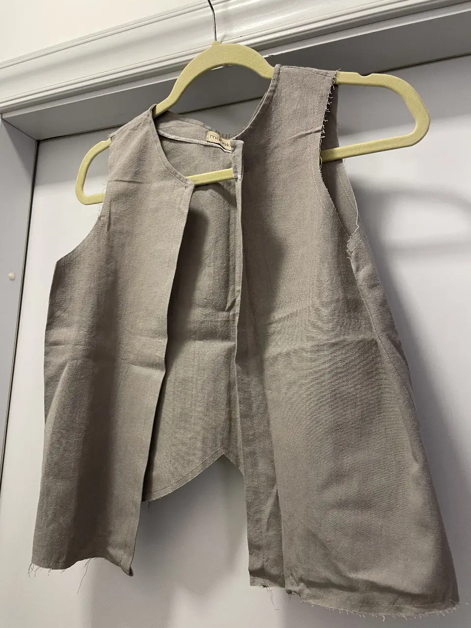 Chaleco de lino gris claro, abierto al frente, con etiqueta de la marca Maneka. Ideal para un look casual y fresco.