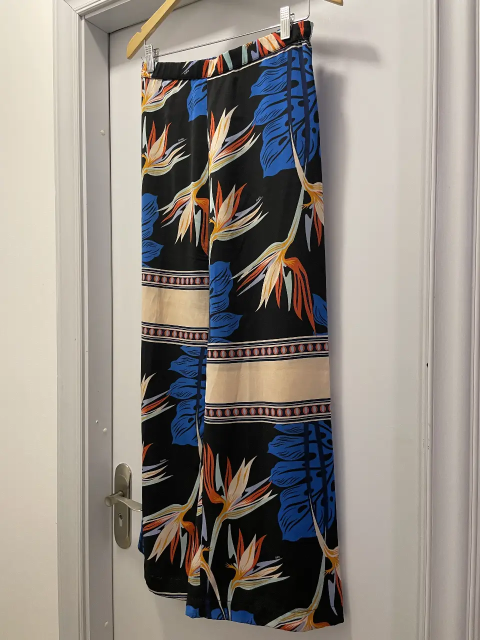 Pantalón de vestir Cher, con estampado floral en tonos naranja, blanco y azul sobre fondo negro. Corte suelto y palazzo, ideal para un look fresco y moderno.