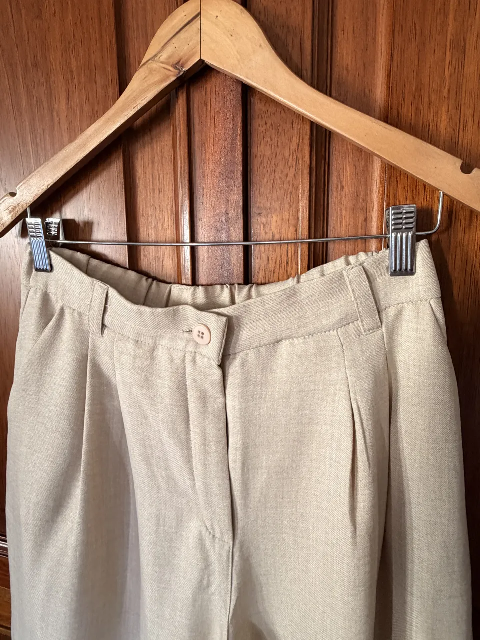 Pantalón de vestir color beige con pinzas delanteras y elástico en la cintura trasera. Posee un botón y cierre en la parte delantera.