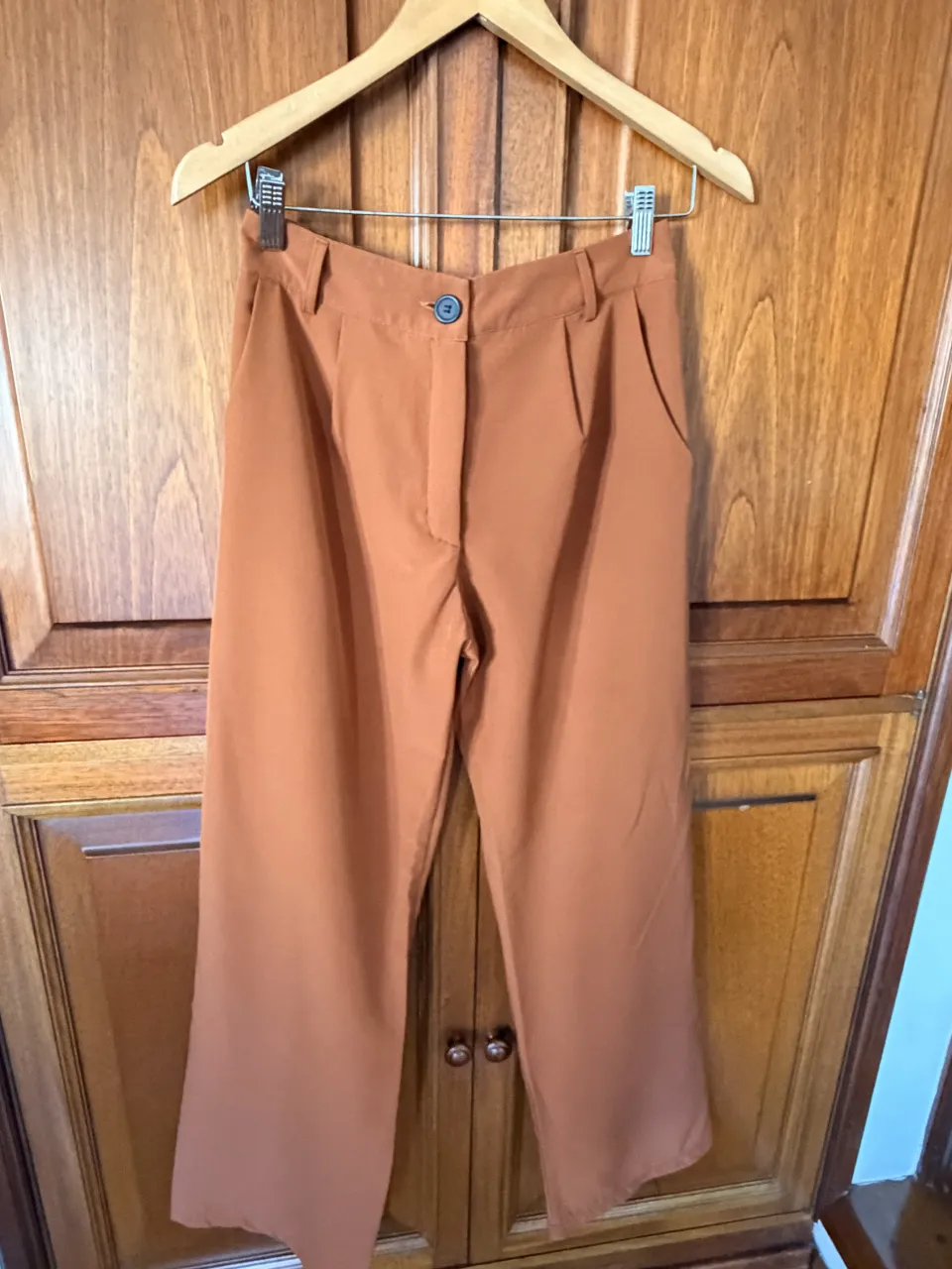 Pantalón color marrón con pinzas delanteras y elástico en la cintura trasera. Tiene un botón y cierre en la parte delantera.