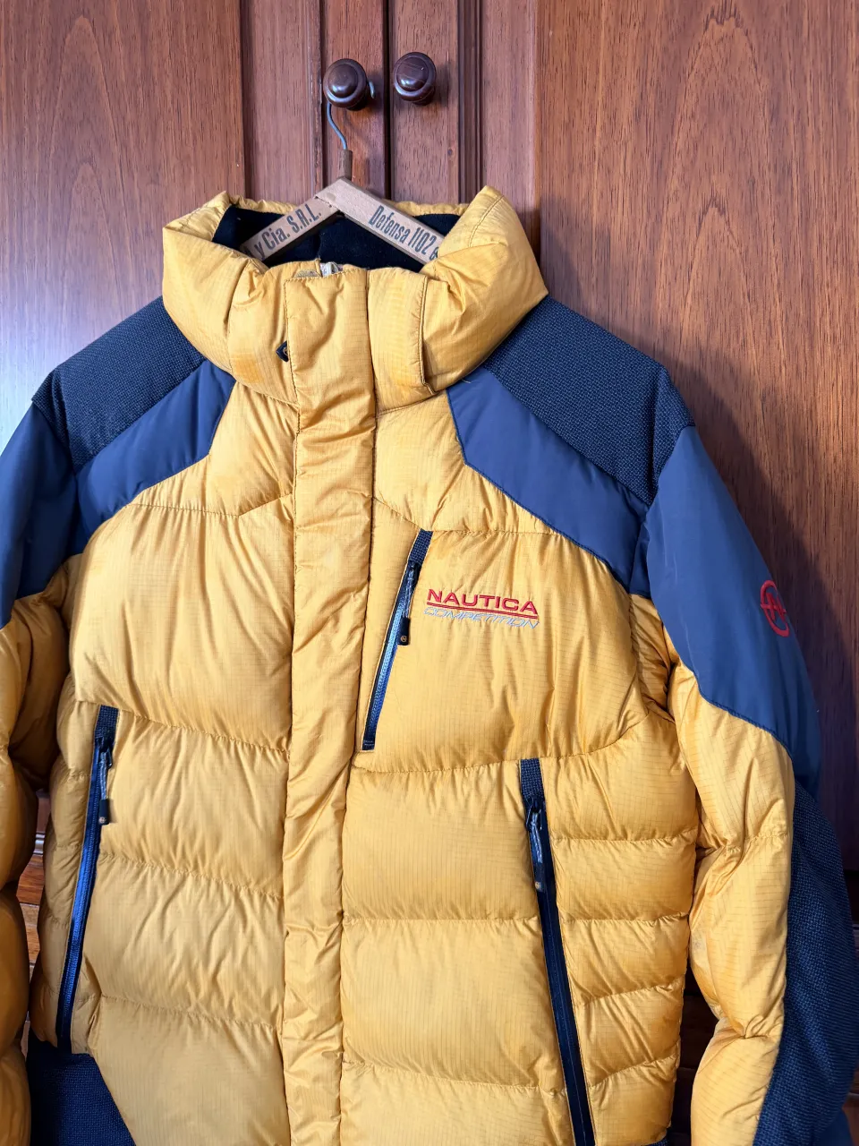 Campera acolchada amarilla y azul marino con detalles grises. Tiene cierre frontal y bolsillos con cierre. Logo de Nautica Competition en el frente y en la manga. 100% pluma.