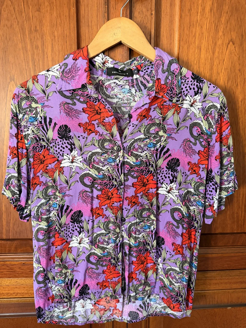 Camisa manga corta con estampado floral y de dragones en tonos lilas, rojos, blancos y negros. Cuello camisero con botones al frente.