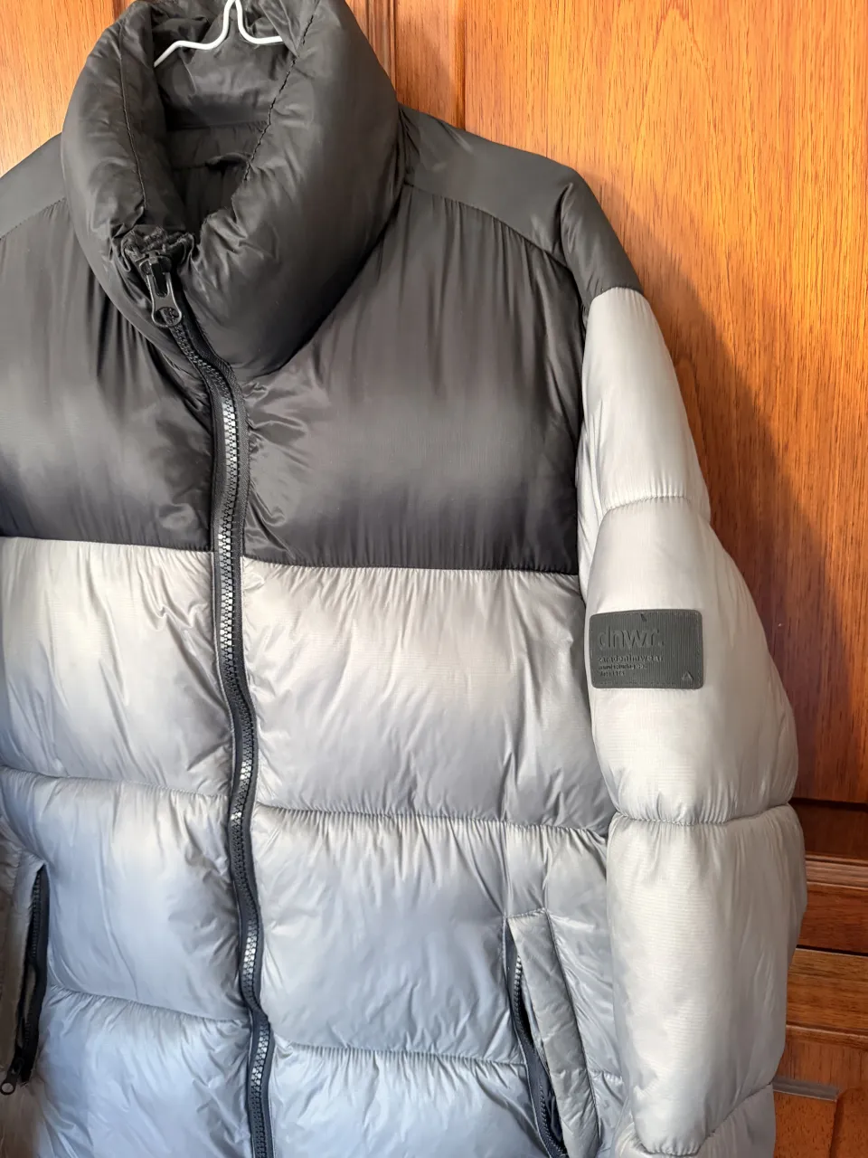 Campera acolchada de dos tonos, gris y negro, con cuello alto y cierre central. Tiene un logo rectangular en la manga izquierda.