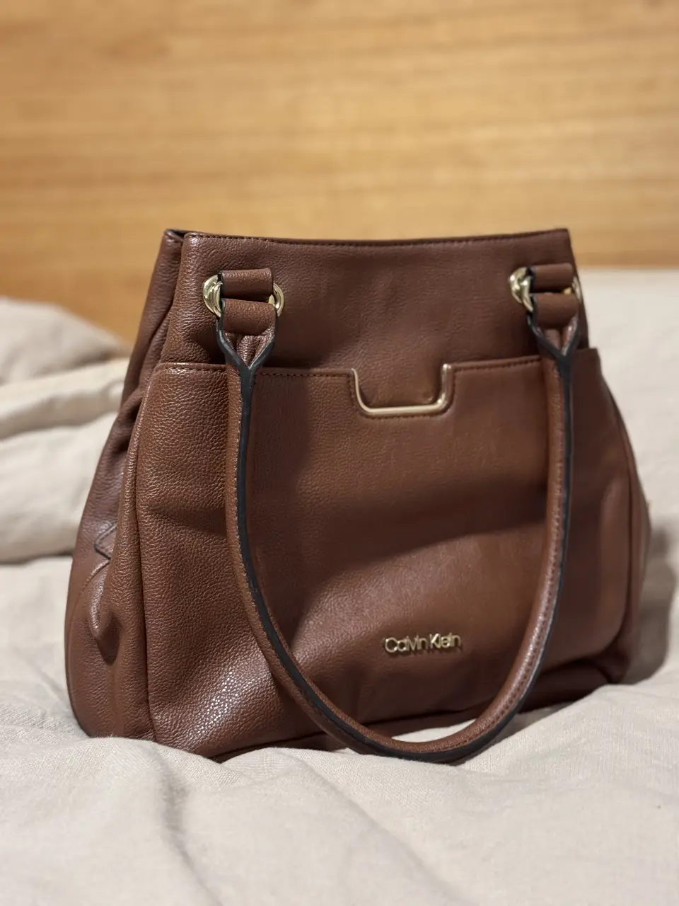 Cartera Calvin Klein marrón, con manijas y detalles dorados. Ideal para llevar tus esenciales con estilo.