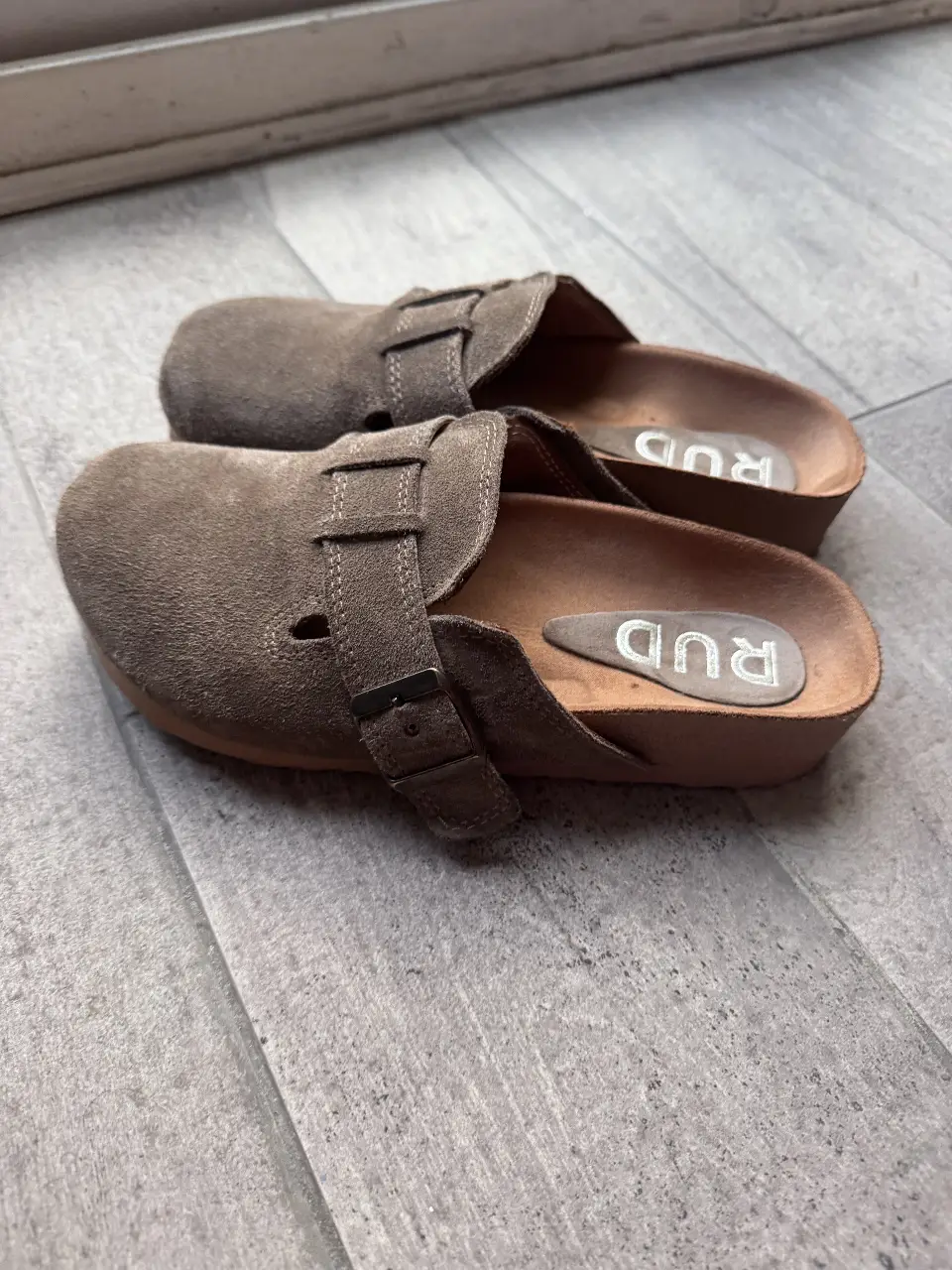 Zuecos RUD de gamuza marrón, con hebilla ajustable y plantilla con logo. Ideales para un look casual y cómodo. tienen 2 usos, para ir literalmente al día de la vuelta de casa, me encantan, pero son 36 y me quedan chicas. mi pie mide 24 cm, son para un 23.7 cm para abajo