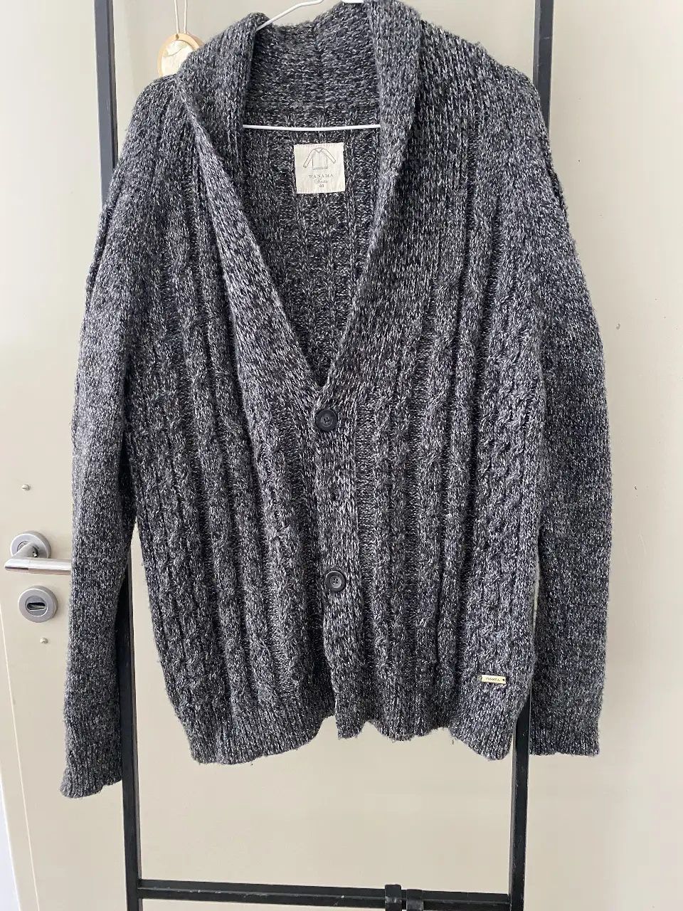 Cardigan de hombre tejido en lana gruesa, color gris con detalles en blanco. Diseño con cuello en V, botones y detalle de trenzas. Ideal para un look casual y abrigado.