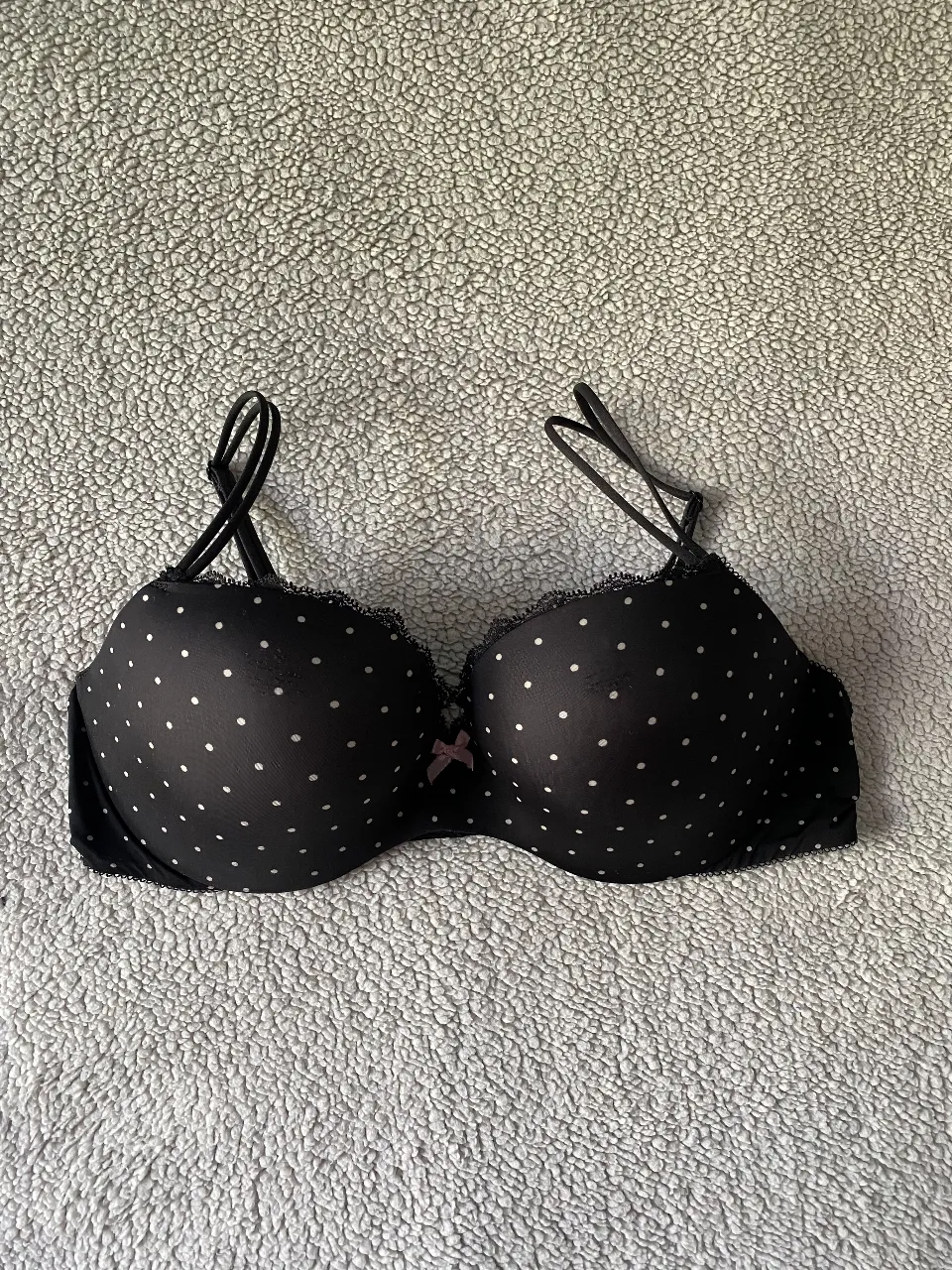 Corpiño negro con lunares blancos y detalle de puntilla en el borde superior. Tiene un pequeño moño rosa en el centro. Ideal para el uso diario o para complementar un look coqueto.