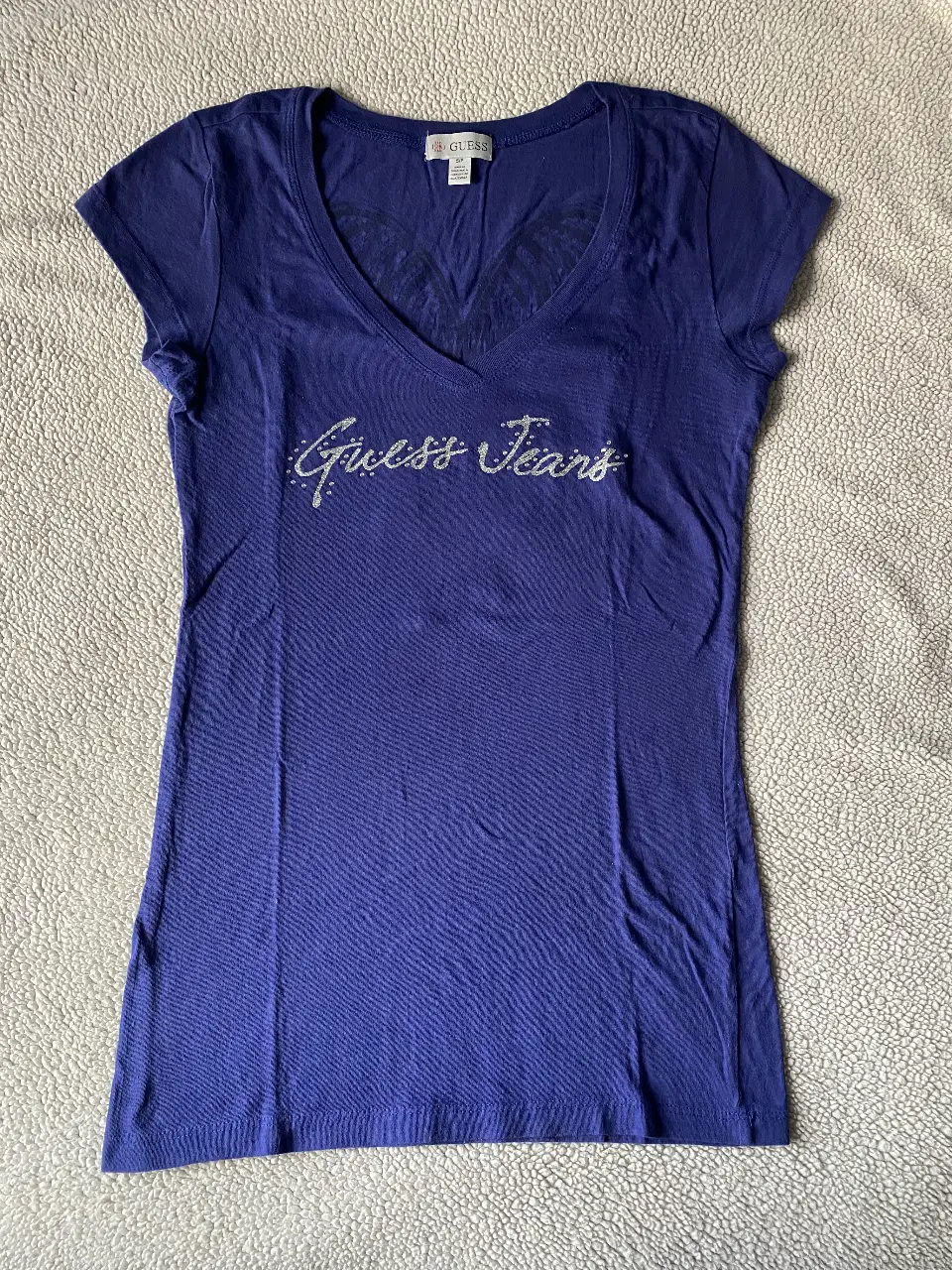 Remera Guess Jeans con estampado de brillos y diseño de alas plateadas en la espalda. Talle S/P.