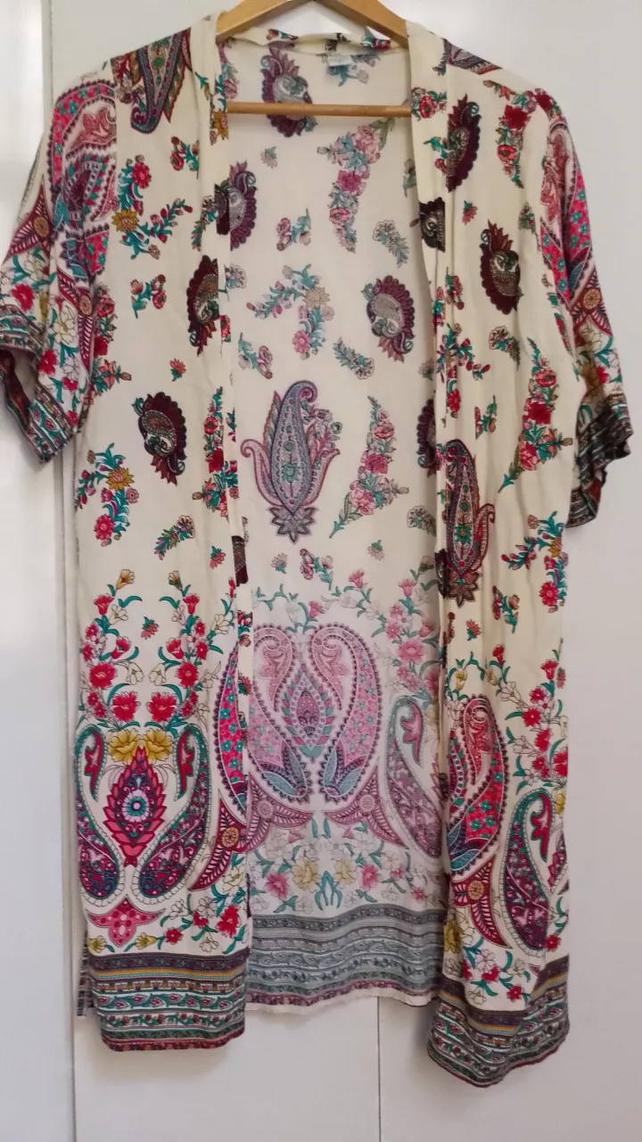 Kimono blanco con estampado de flores y paisleys en tonos rosas, rojos, verdes y azules. Posee mangas cortas y terminación en flecos en la parte inferior.