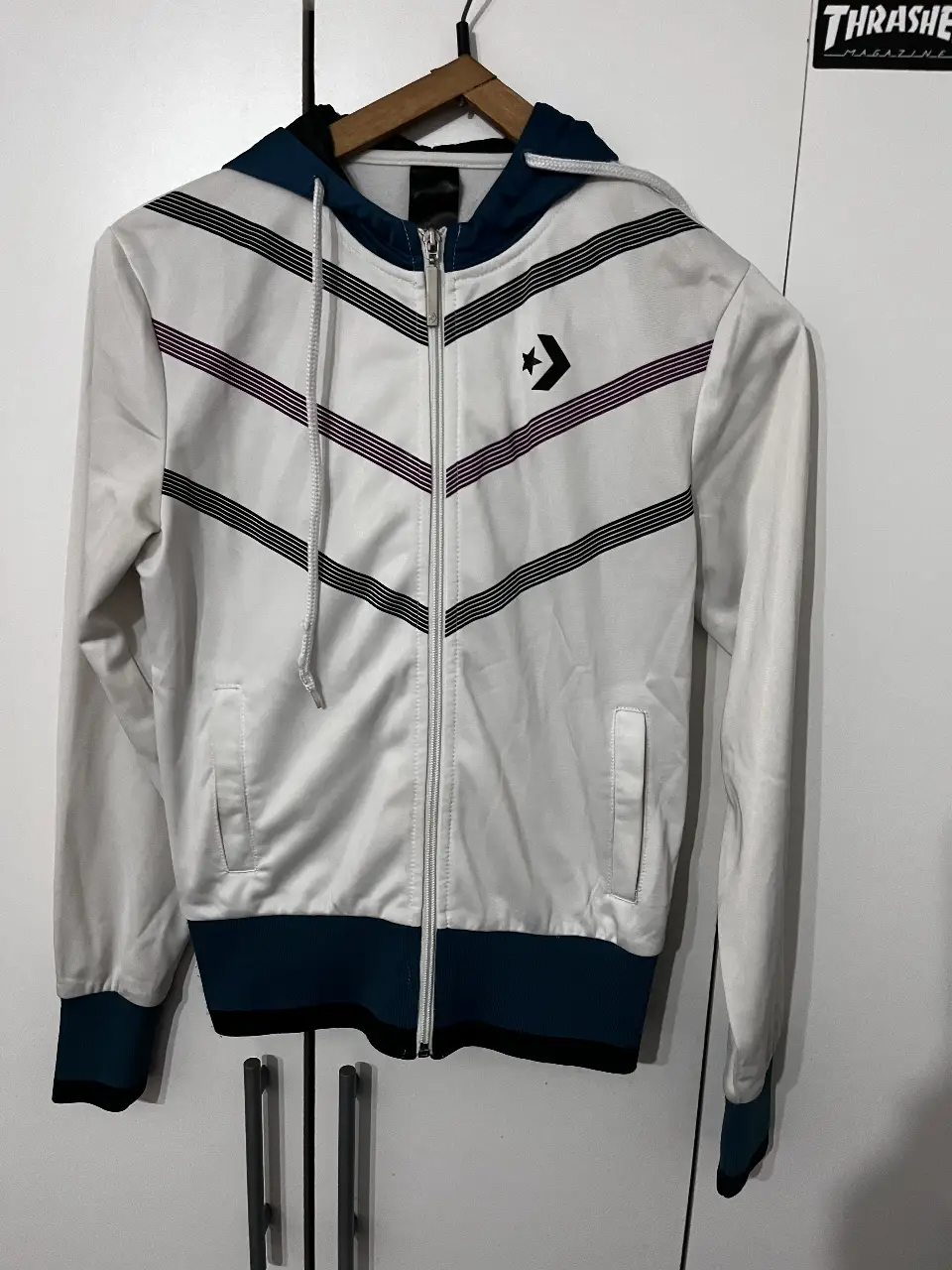Campera deportiva Converse blanca con detalles en azul y negro. Diseño con cierre frontal, capucha y el logo de la marca en el pecho. Ideal para un look casual y cómodo.