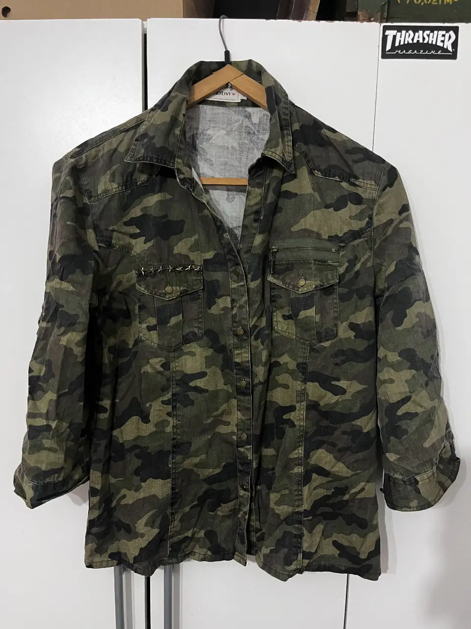 Camisa de hombre con estampado camuflado en tonos verdes y negros. Corte clásico con mangas 3/4, bolsillos delanteros y detalles metálicos. Ideal para un look casual y moderno.