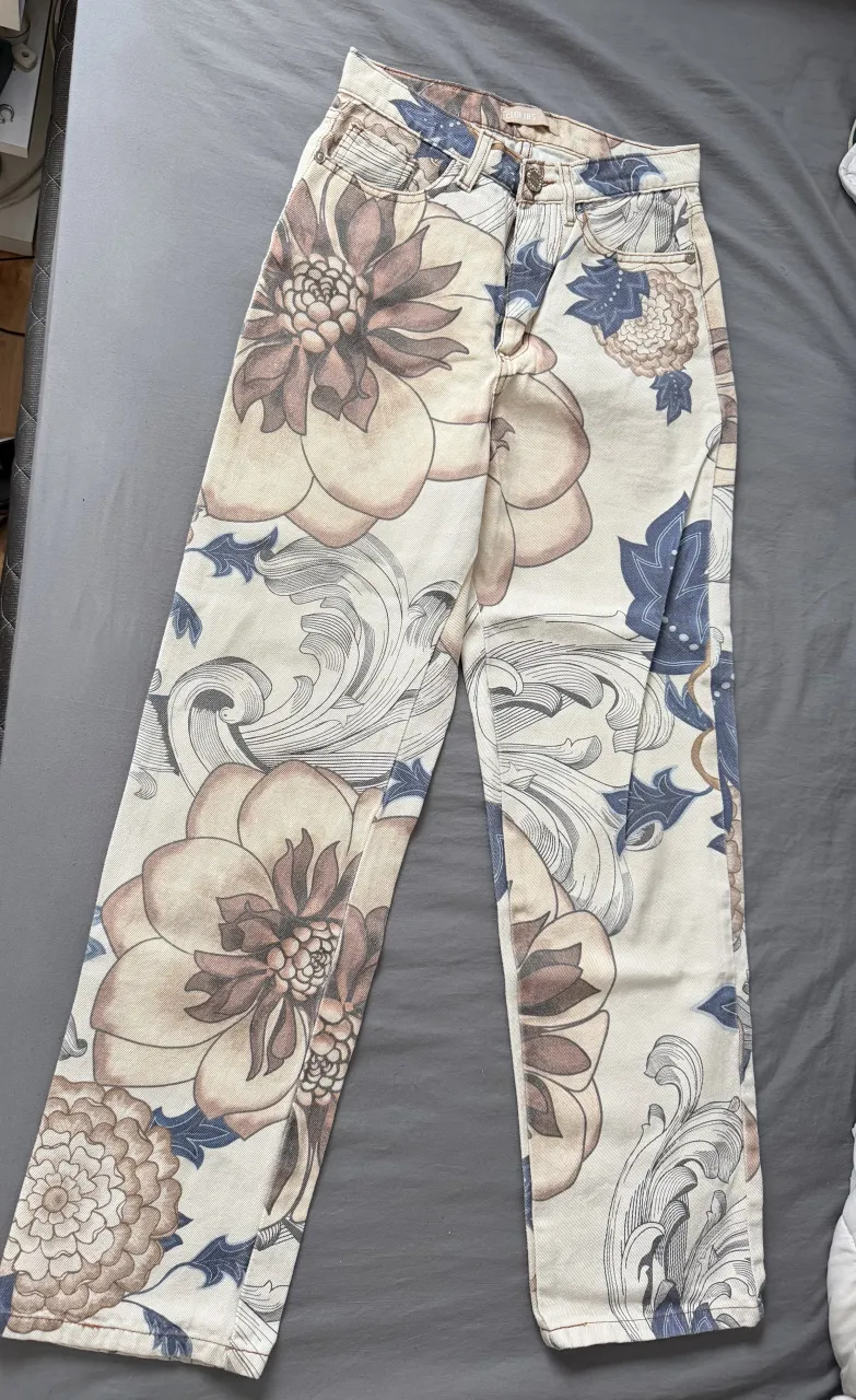 Jeans de tiro alto con estampado floral grande en tonos beige, marrón y azul sobre fondo blanco. Cuentan con cuatro bolsillos y cierre.