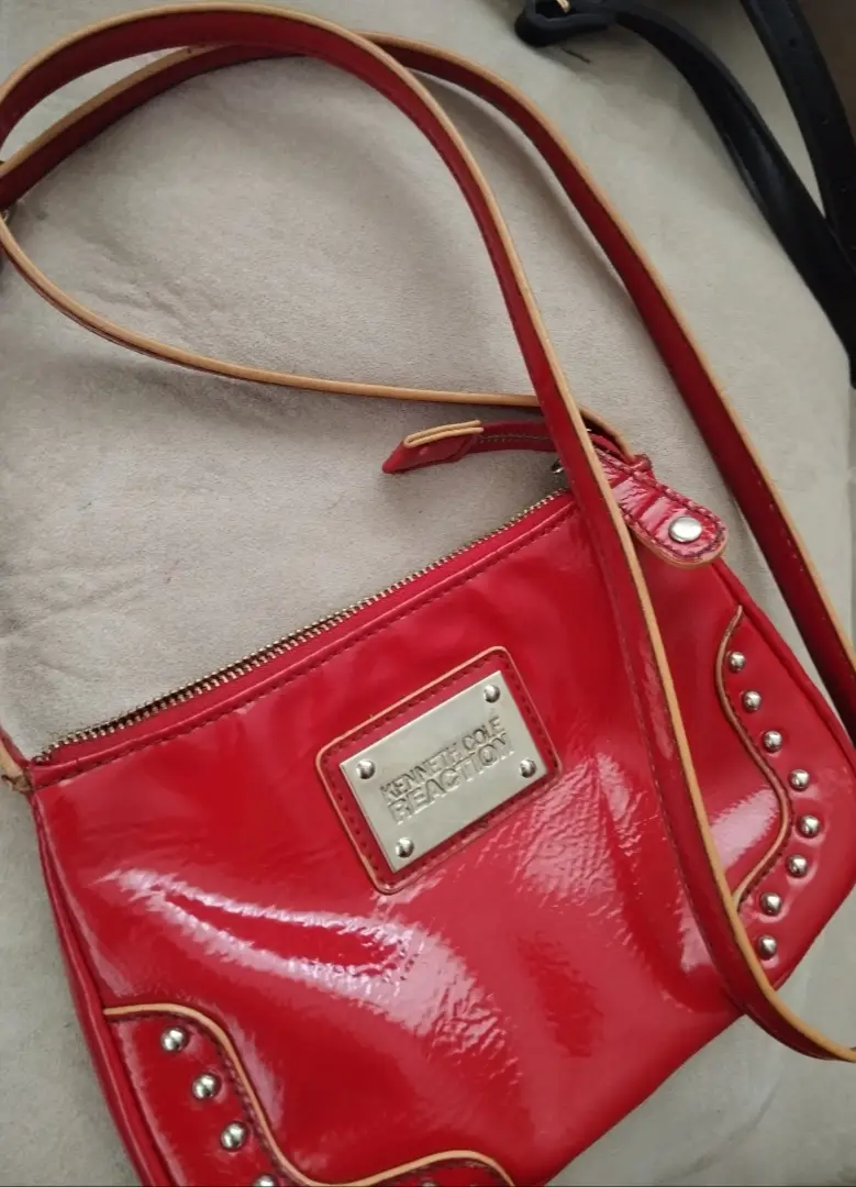 Bandolera roja de charol con detalles de tachas plateadas y placa con el logo de la marca. Ideal para un look moderno