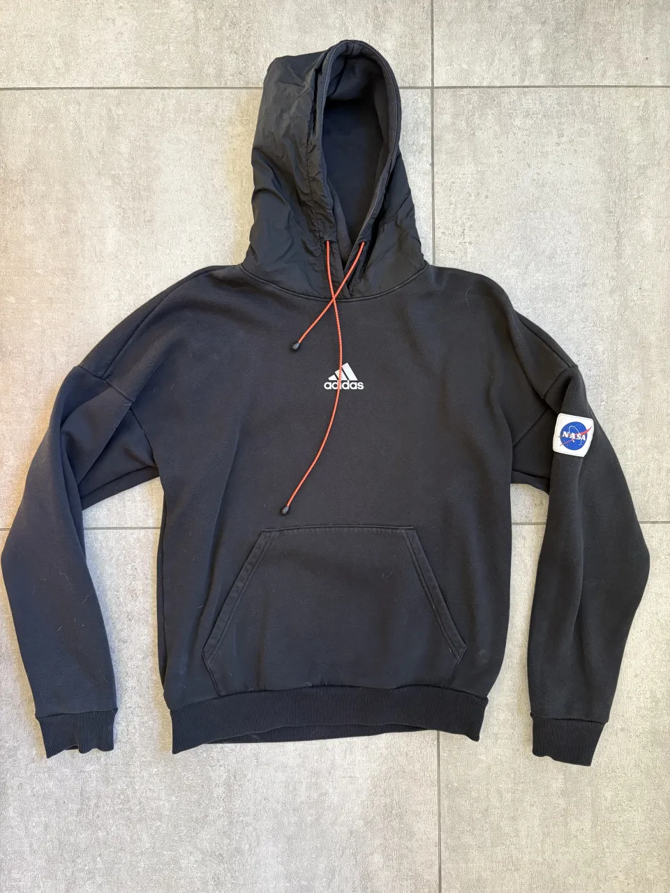 Buzo negro con capucha, de Adidas y NASA. Tiene el logo de Adidas en el frente y un parche de la NASA en la manga izquierda. En la espalda tiene un estampado triangular con los logos de Adidas y NASA. Cordones naranjas.  La capucha es como una tela medio técnica, tipo rompevientos.Talle 1 de hombre o 2 de mujer.