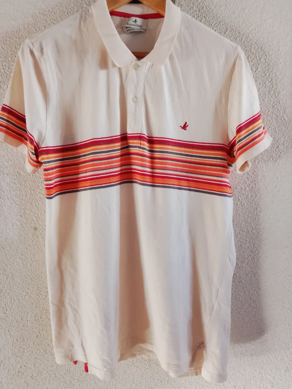 Remera polo blanca con cuello y mangas de tejido a rayas horizontales en tonos naranja, rojo y gris. Presenta un pequeño bordado de un pájaro en color rojo en el frente. Tiene dos botones en el cuello.