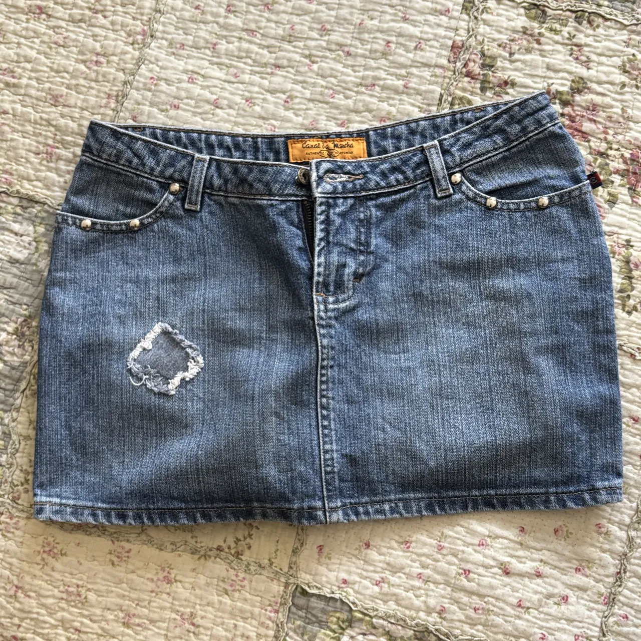 Minifalda de jean azul tiro bajo con detalles de tachas y un parche decorativo. Ideal para un look casual y moderno. Perfecta para combinar con remeras, tops o camisas.
